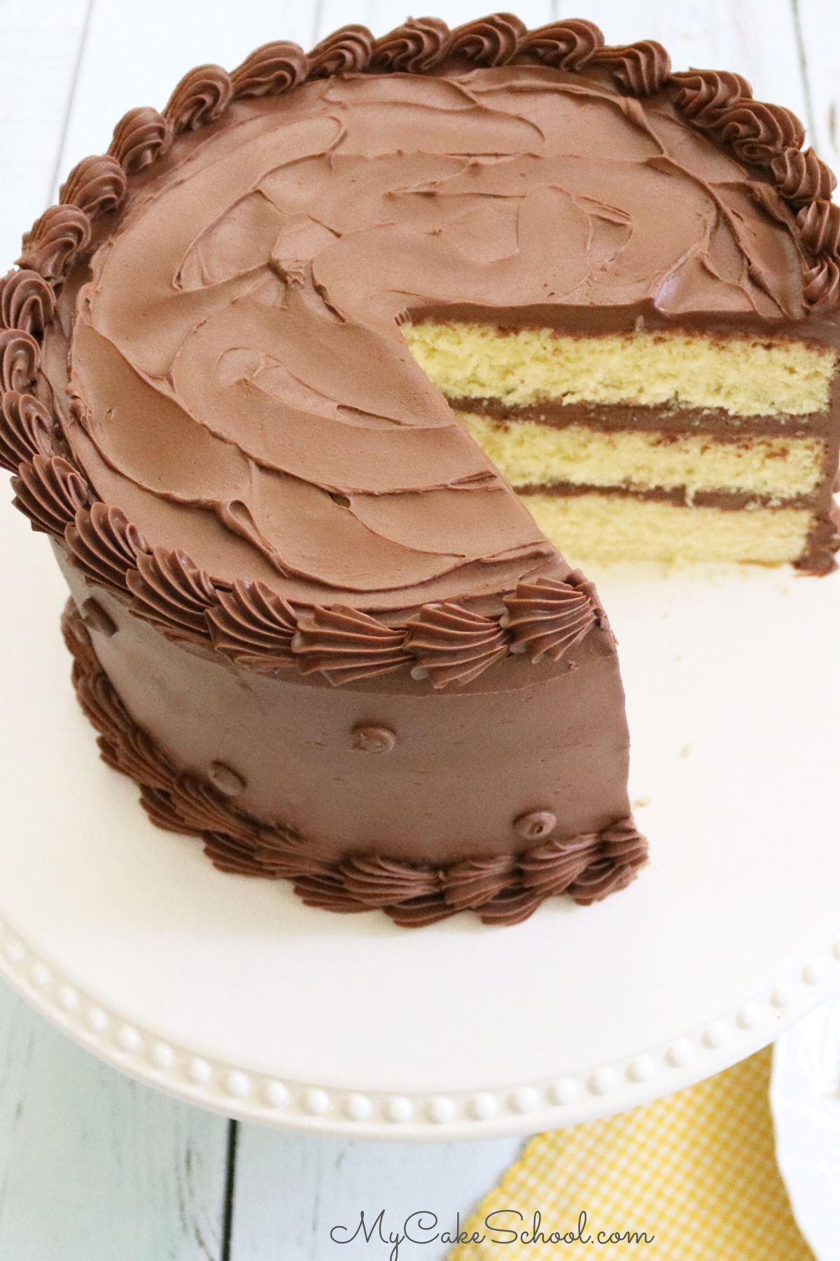 Sliced yellow cake from scratch on a white pedestal, frosted in chocolate buttercream.