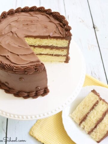 Sliced yellow cake with chocolate frosting on white pedestal with a plated slice next to it.