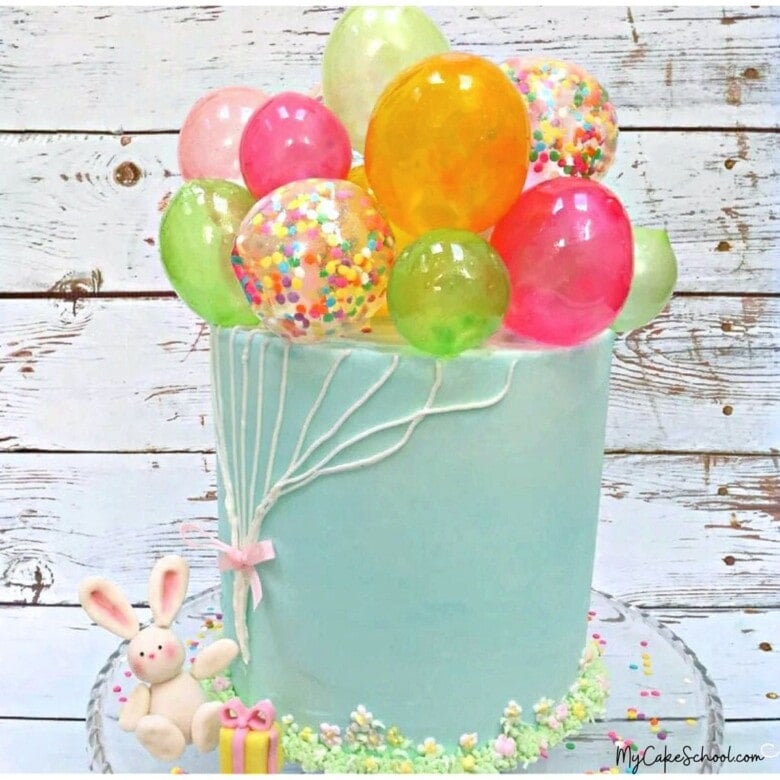 Gelatin Balloons Cake. Blue frosted cake with colorful gelatin balloons on top, and fondant bunny holding the buttercream "strings".
