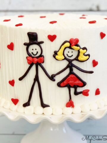 Cake on pedestal with chocolate stick figure decoration on front.