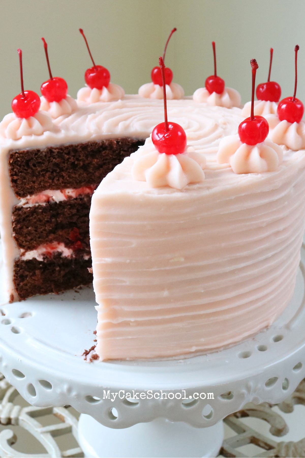 Cherry Cherry Cake, with cherry cream cheese frosting, topped with maraschino cherries, on white pedestal.