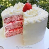 Sliced Strawberry Cake with white chocolate frosting on a white pedestal, topped with white chocolate curls and a strawberry.