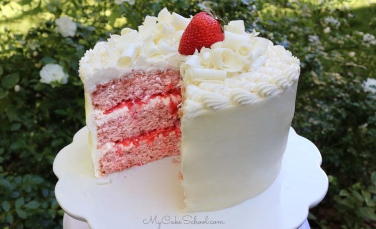 Sliced Strawberry Cake with white chocolate frosting on a white pedestal, topped with white chocolate curls and a strawberry.