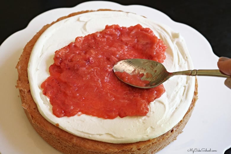 Spreading cooked strawberry filling on top of a layer of strawberry sour cream cake spread with white chocolate frosting.