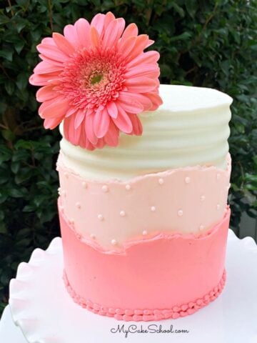 Cake with a layered buttercream cake design (white frosting, peach frosting, and pink frosting) on a white pedestal. Topped with pink gerbera daisy.