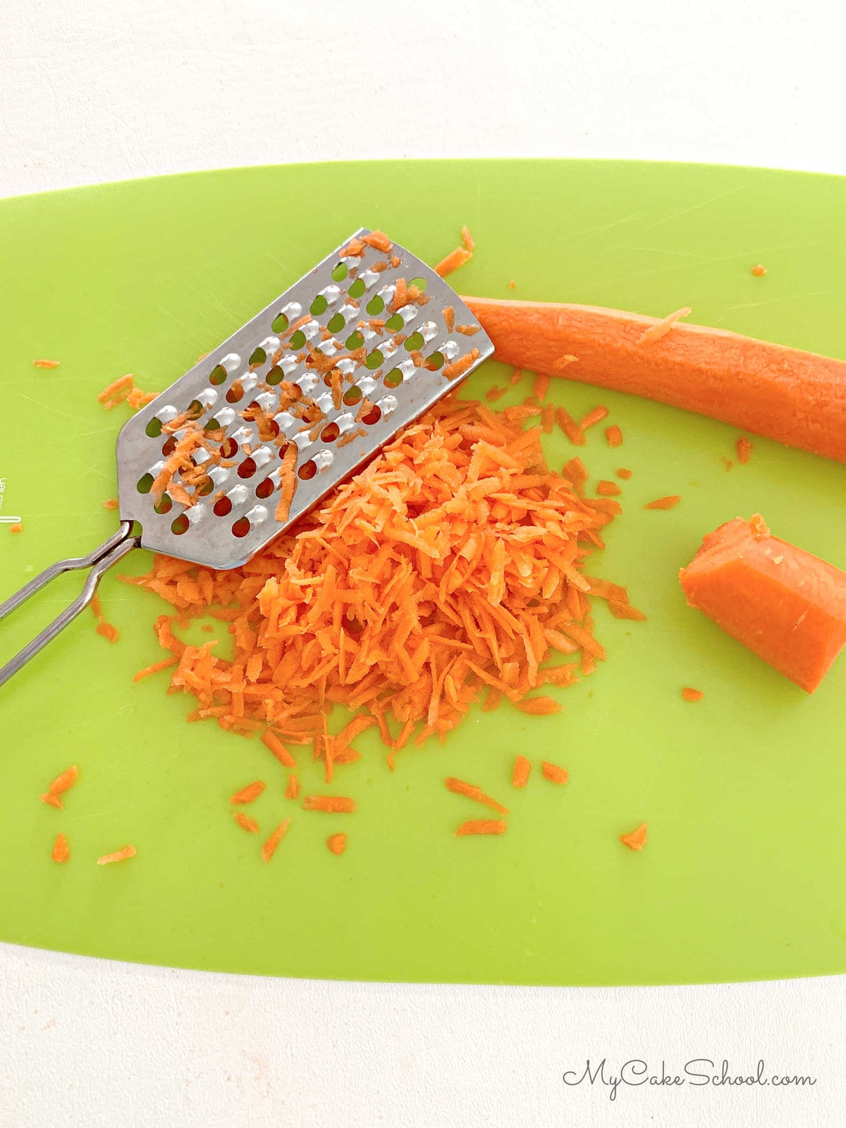A mound of grated carrots on a cutting board, next to grater and whole carrots.