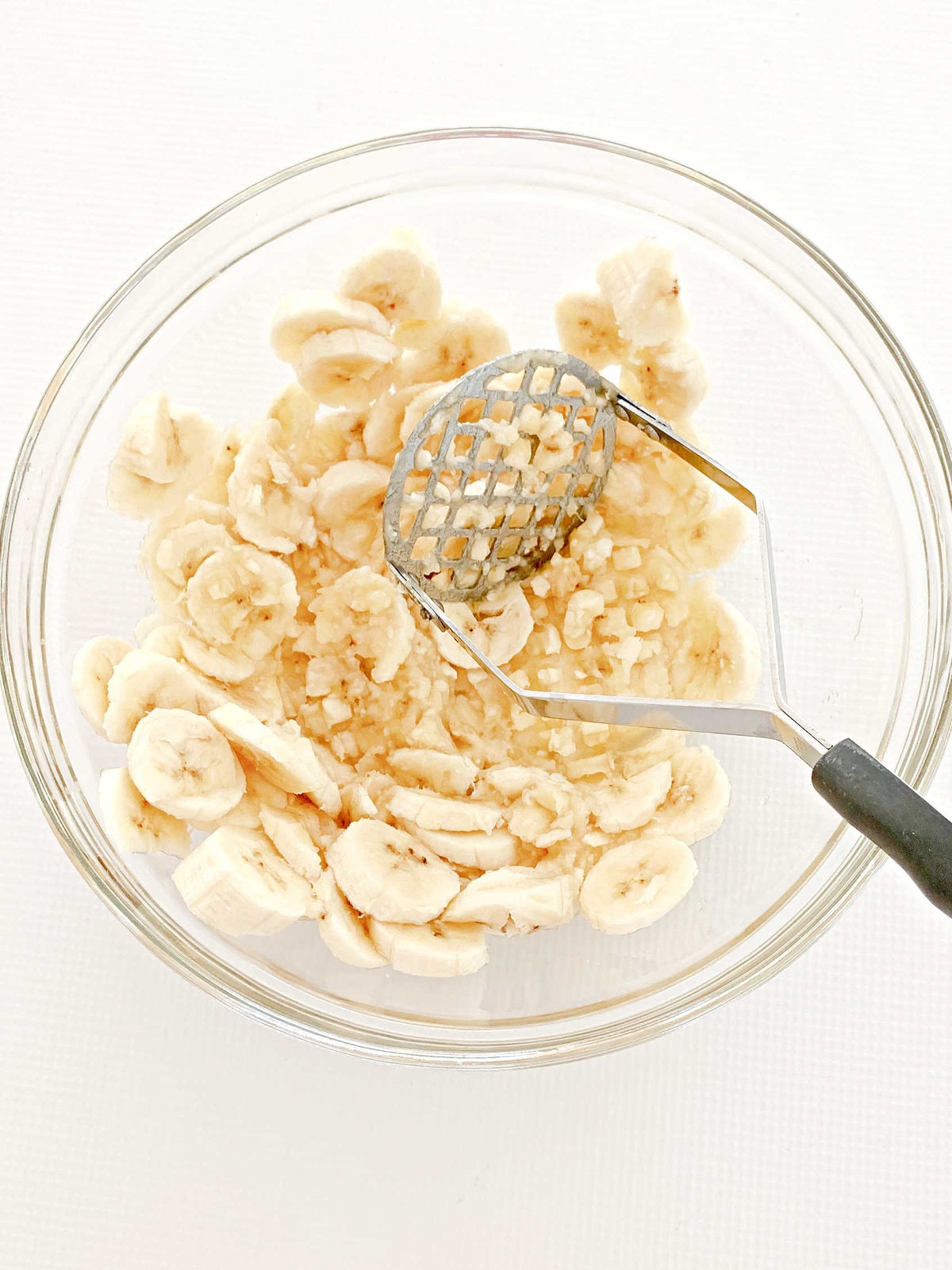 Sliced bananas and potato masher in bowl