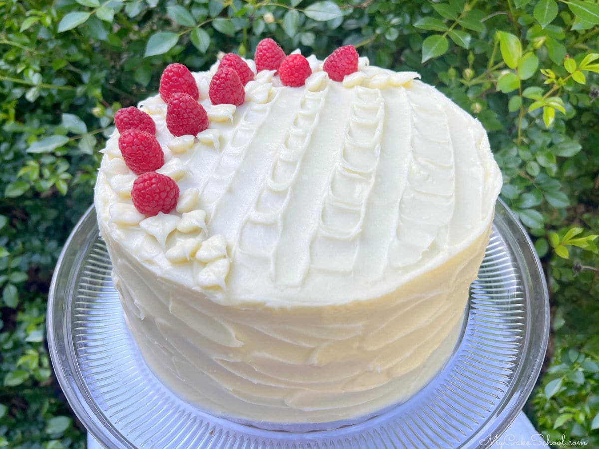 Decorated Raspberry Cake on pedestal. It's frosted with cream cheese frosting and topped with fresh raspberries.