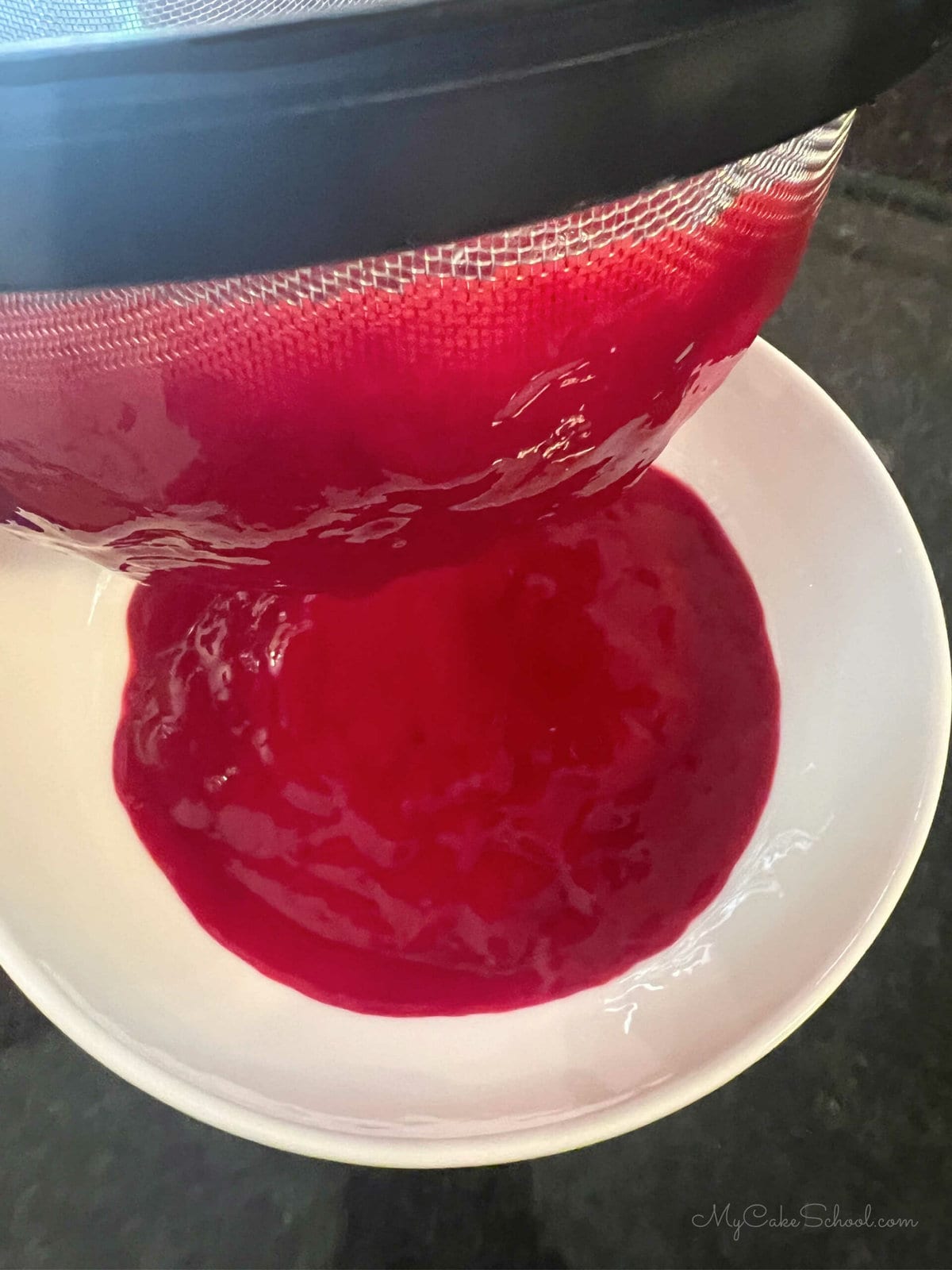 Straining raspberry puree through a fine strainer and into a white bowl, removing seeds.  