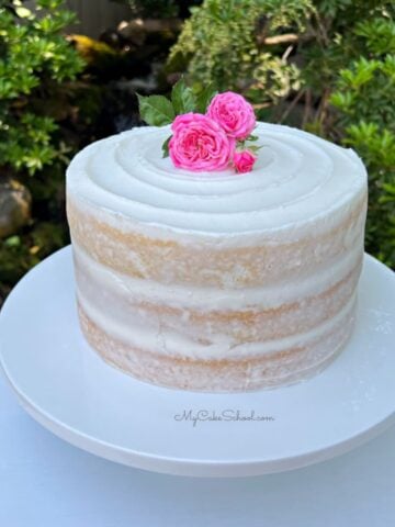Semi-naked cake on a white cake pedestal, topped with fresh pink roses