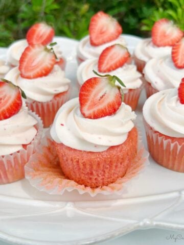 Frosted strawberry cupcakes on a platter, topped with strawberry halves.