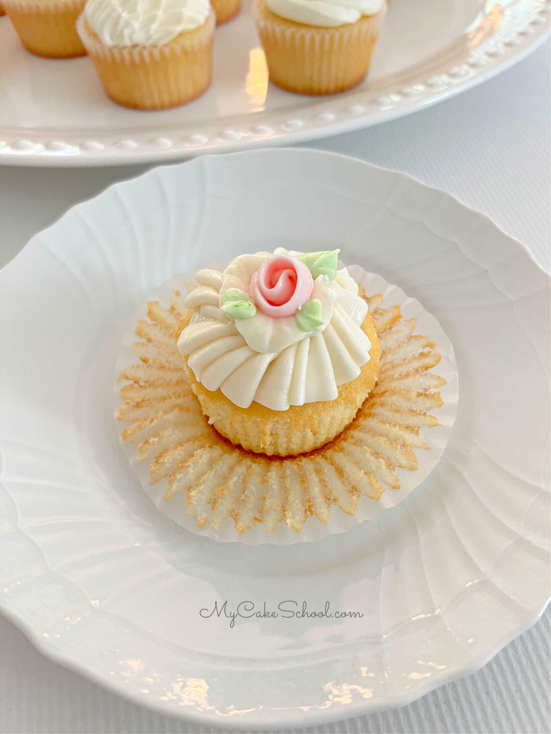 Unwrapped vanilla cupcake on a plate, frosted with buttercream ruffles and topped with a pink buttercream ribbon rose.