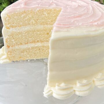 Sliced homemade white cake on glass pedestal. White frosting with light pink frosting swirls on top.