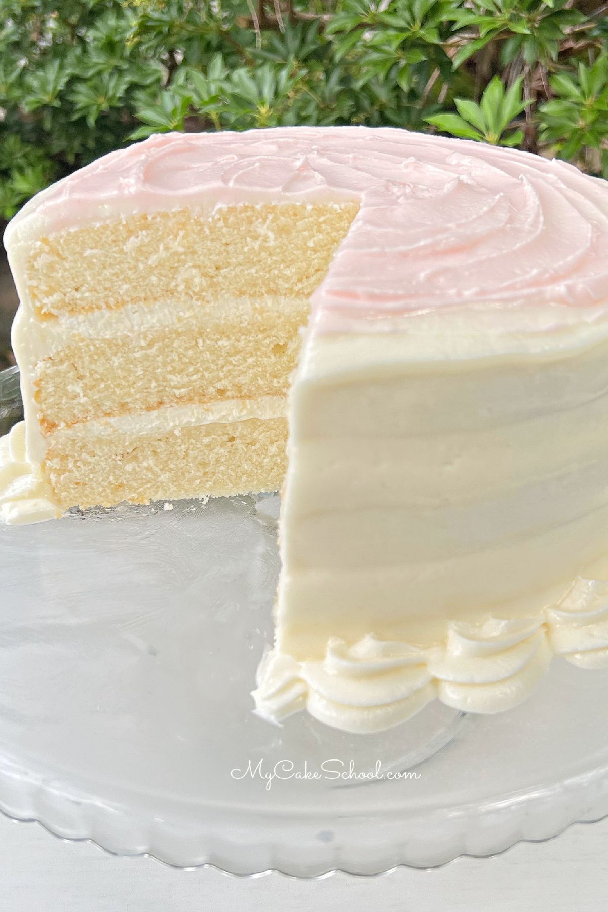 Sliced homemade white cake on glass pedestal. White frosting with light pink frosting swirls on top.
