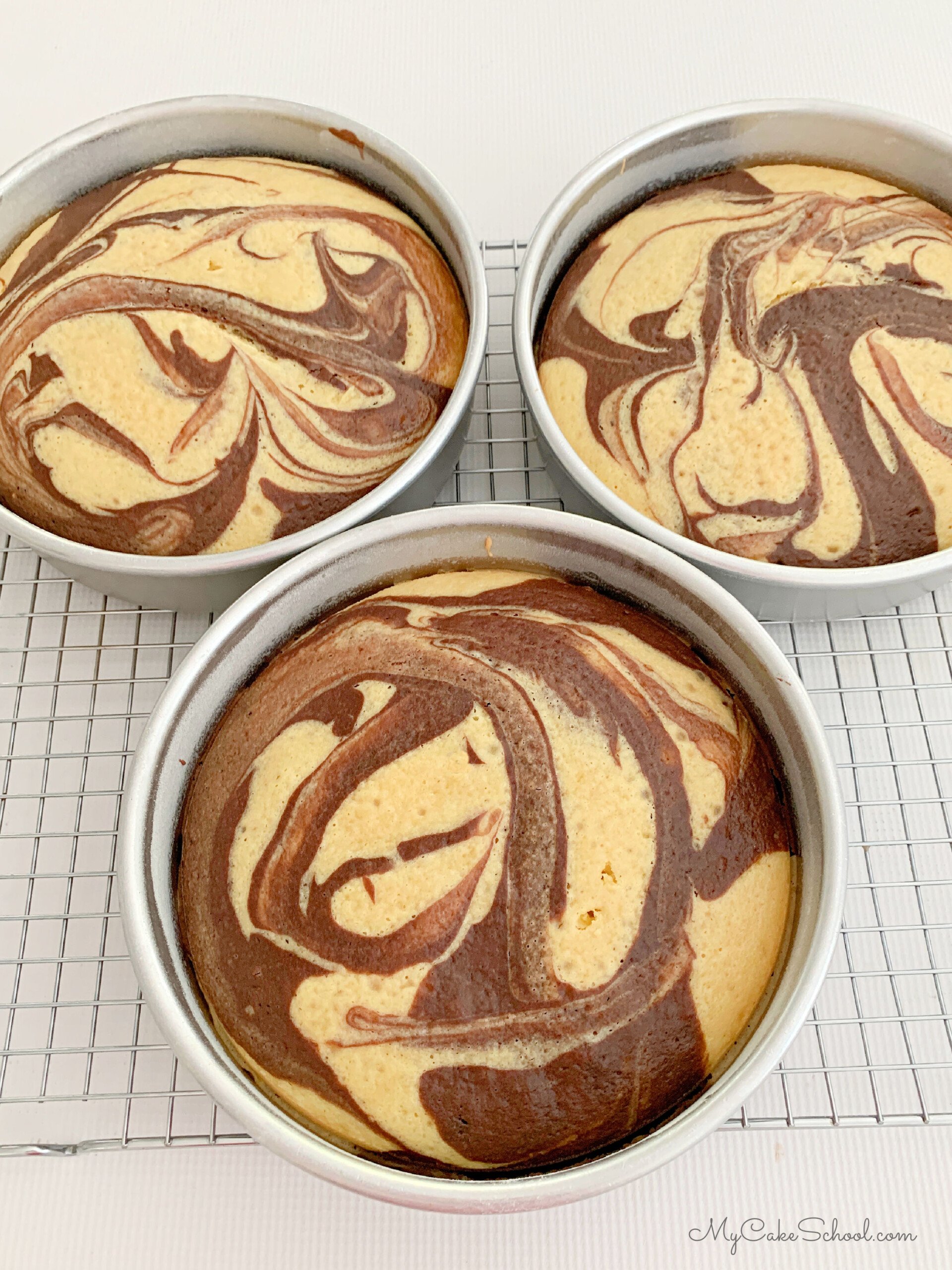 Three baked marble cakes in 8 inch cake pans, cooling on a wire rack.