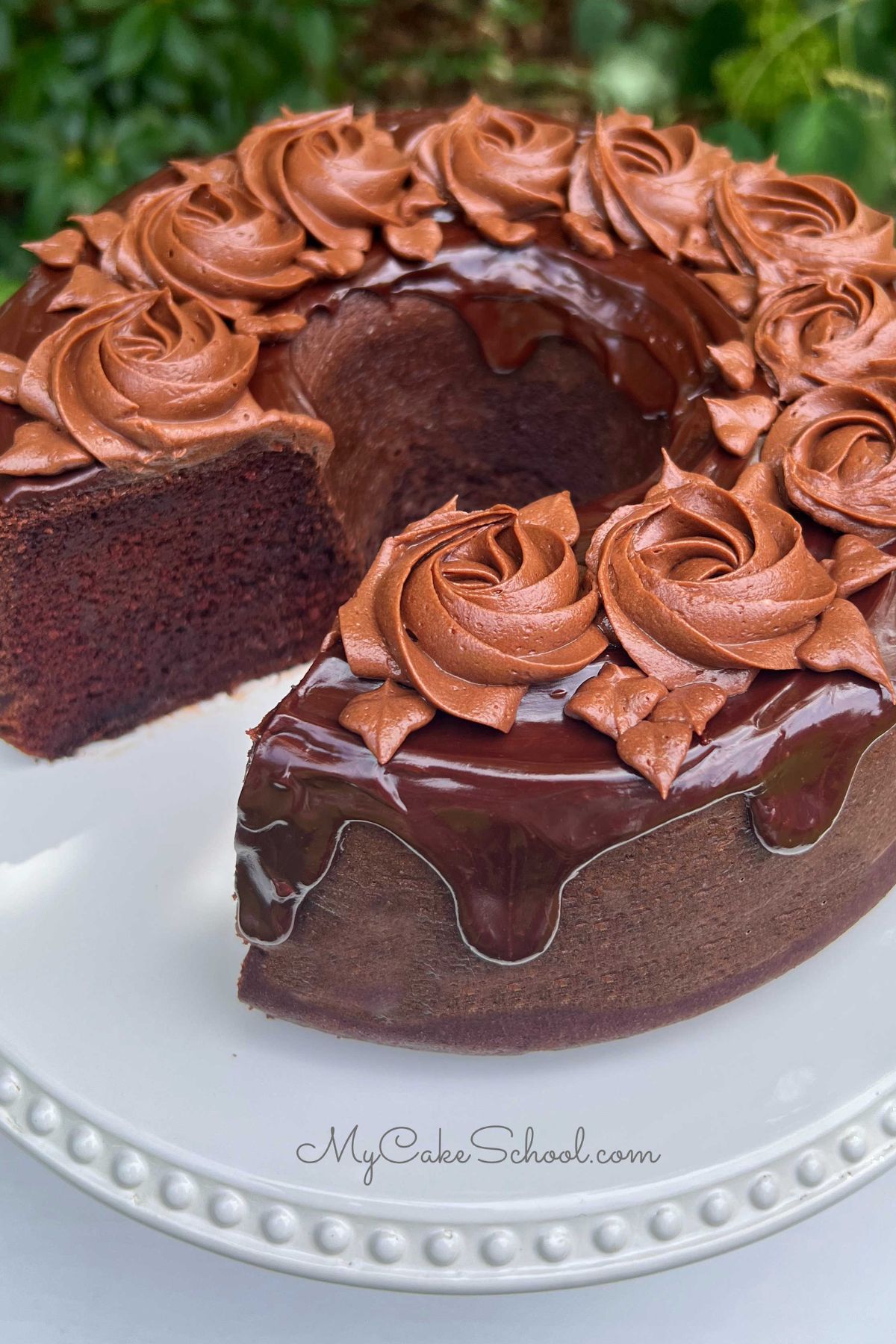 Chocolate Bundt Cake topped with chocolate glaze and chocolate buttercream rosettes, on a white pedestal.