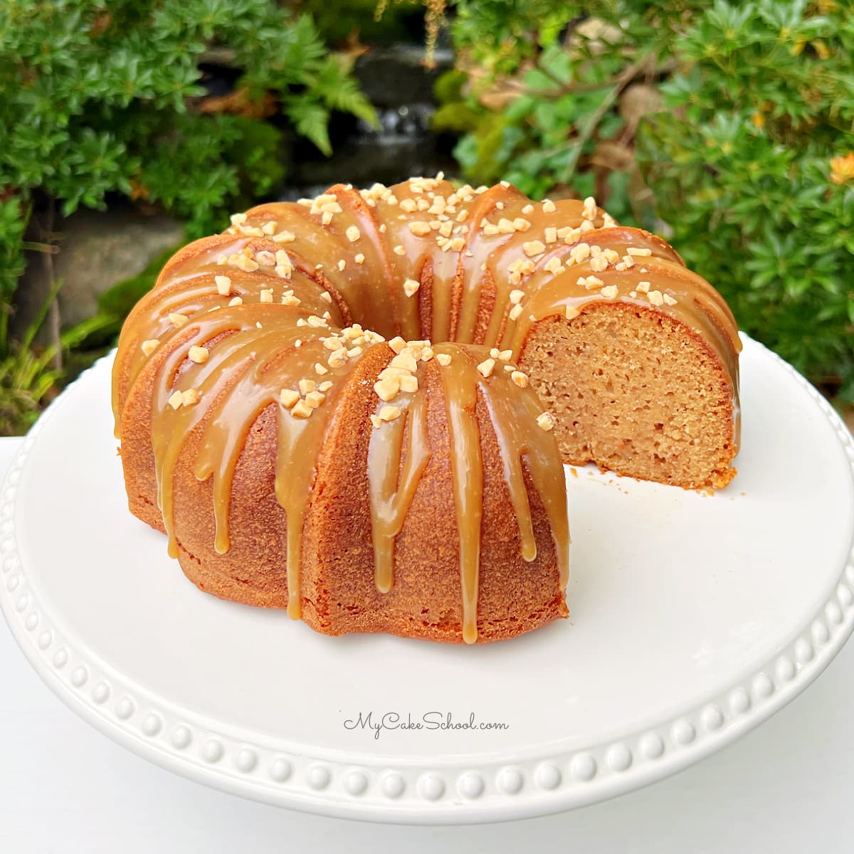 Applesauce cake, sliced, on a pedestal. It is topped with a caramel glaze and toffee bits.