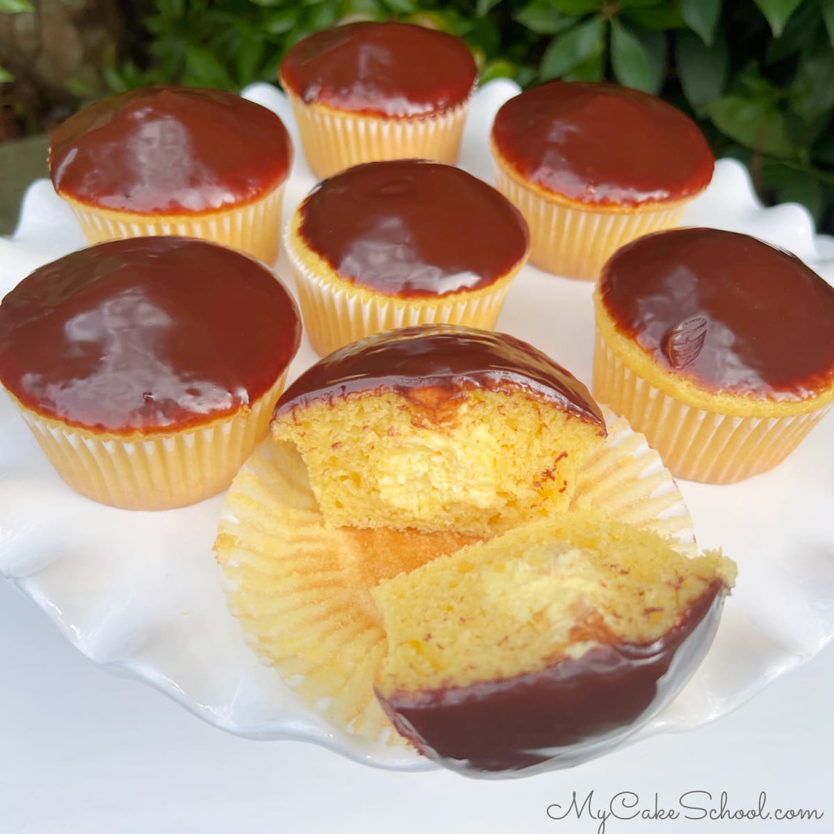 Boston Cream Cupcakes on a cake pedestal. The first cupcake is split in half to reveal the cream filling.