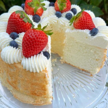 Sliced Angel Food Cake on a pedestal, topped with whipped cream, strawberries, and blueberries.