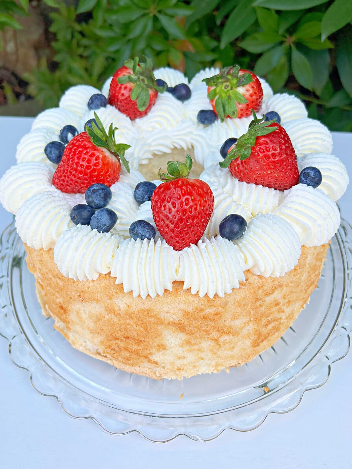 Angel Food Cake on a cake pedestal with whipped cream, strawberries, and blueberries.