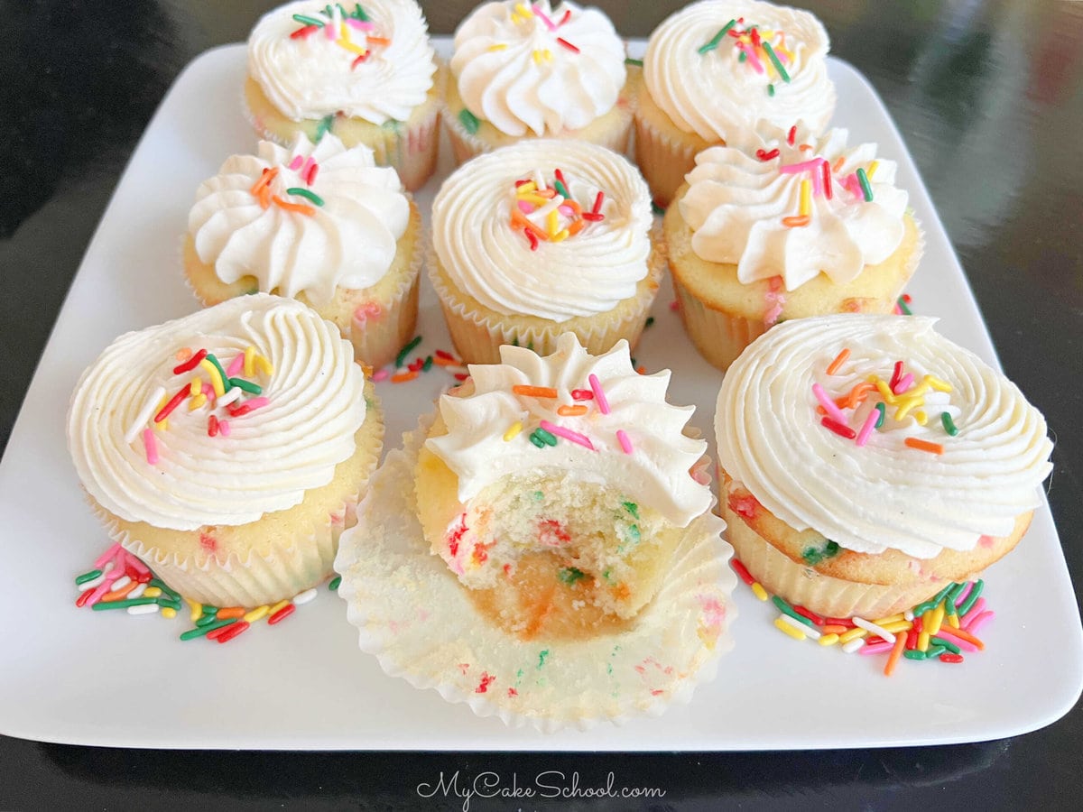 Funfetti Cupcakes on a cake platter, one cupcake has a bite out of it so that you can see the colorful speckled inside.