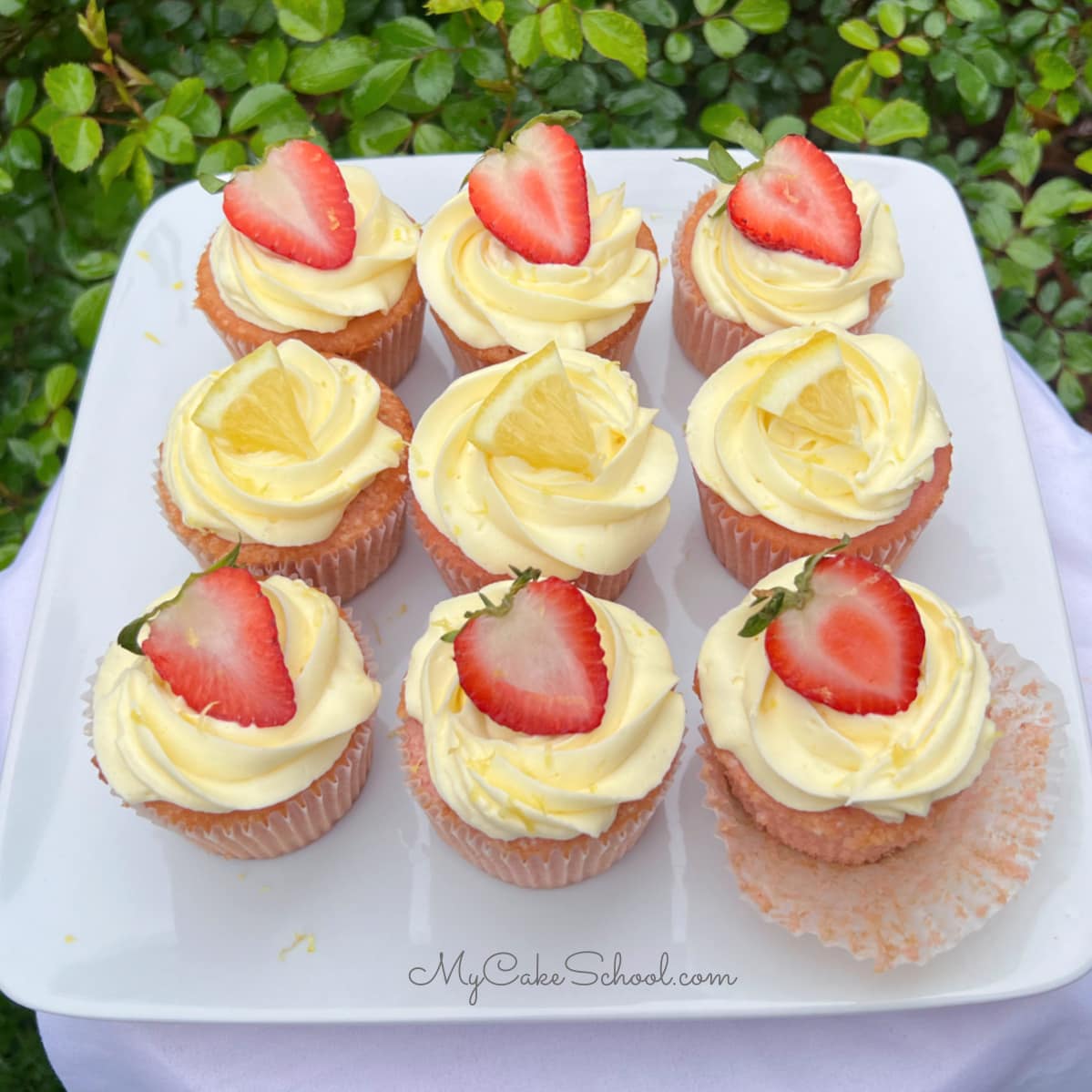 Strawberry lemonade cupcakes on a platter, topped with swirls of lemon whipped cream cheese frosting and topped with sliced strawberries and lemons.