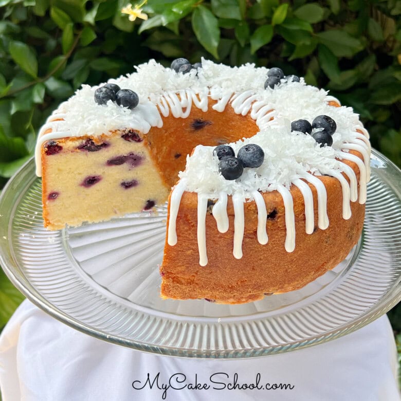 Blueberry Coconut Pound Cake, sliced, on a pedestal. It is topped with a coconut glaze, flaked coconut, and fresh blueberries.